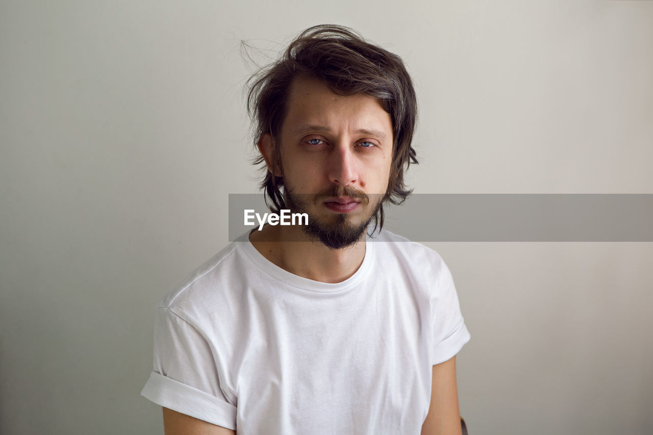 Guy with a beard in a white t-shirt and blue jeans against the white wall of the house