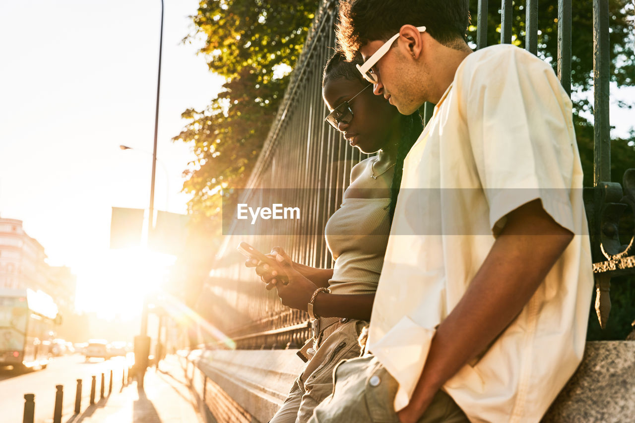 Low angle of diverse couple leaning on park fence and browsing cellphone together while spending time on city street at sunset