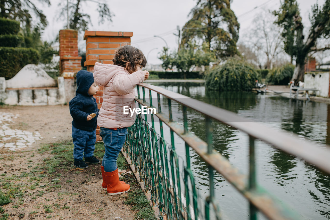 Side view of siblings standing by railing over lake against trees