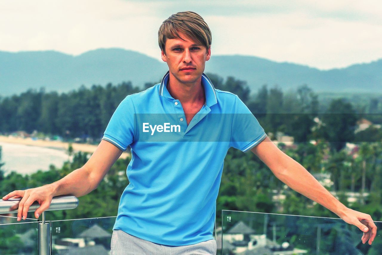 Young man looking away while standing on railing against mountain