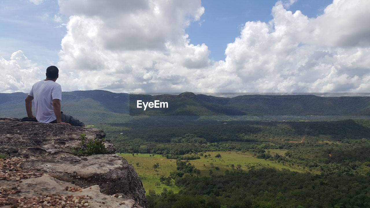 Rear view of man looking at view while sitting on mountain against cloudy sky