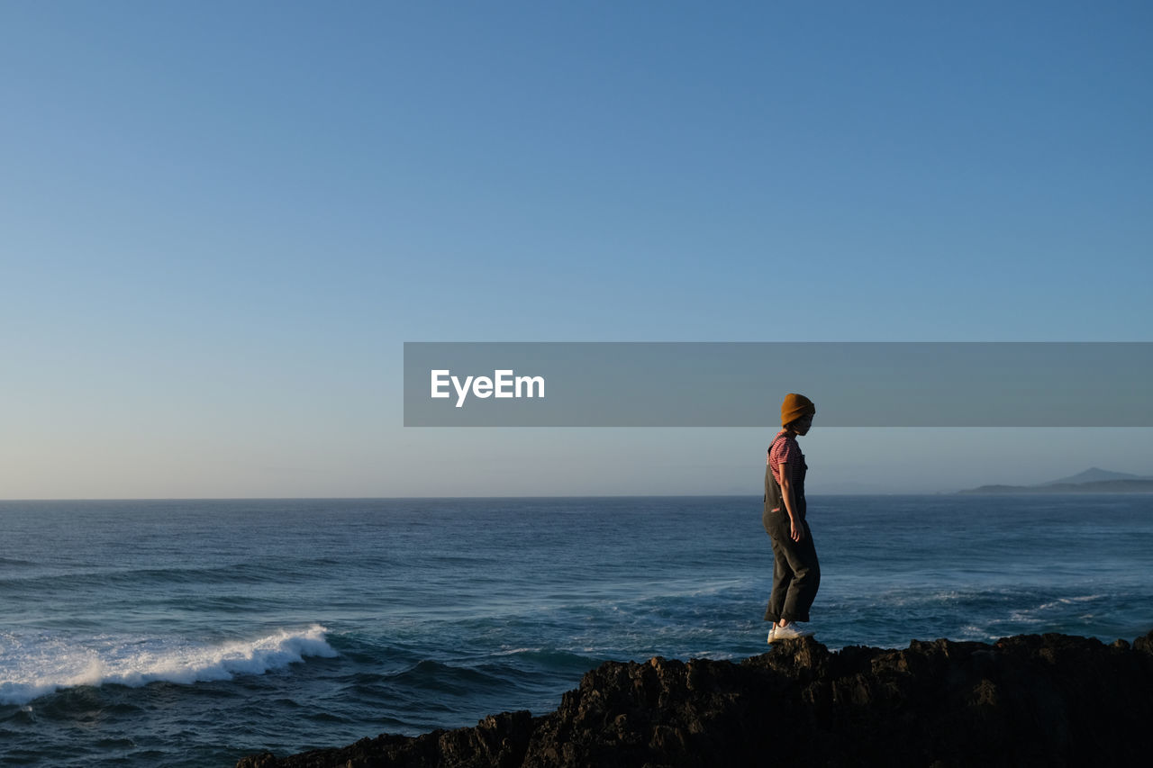 horizon, sky, sea, water, one person, ocean, standing, beauty in nature, horizon over water, nature, land, clear sky, scenics - nature, full length, beach, coast, shore, copy space, adult, tranquility, leisure activity, blue, wave, body of water, rock, holiday, solitude, side view, vacation, lifestyles, trip, tranquil scene, idyllic, looking at view, cliff, outdoors, motion, men, day, sunset, travel, non-urban scene, sports, young adult, activity, dusk, sunlight, travel destinations, terrain, looking, sunny