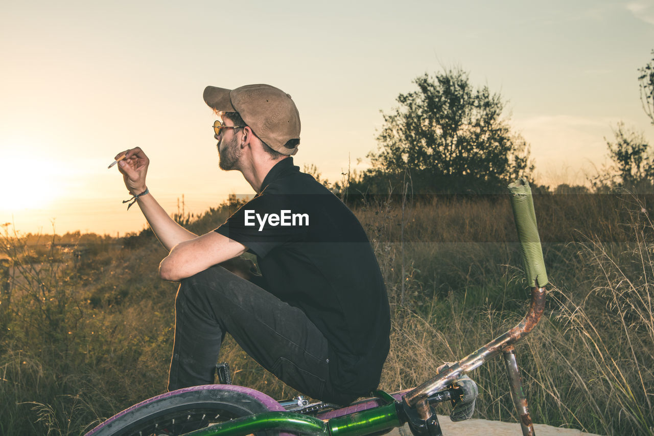 Man sitting with bicycle on field against sky during sunset