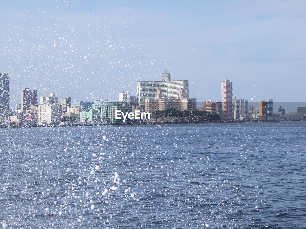 Surf splashing over sea by cityscape against sky