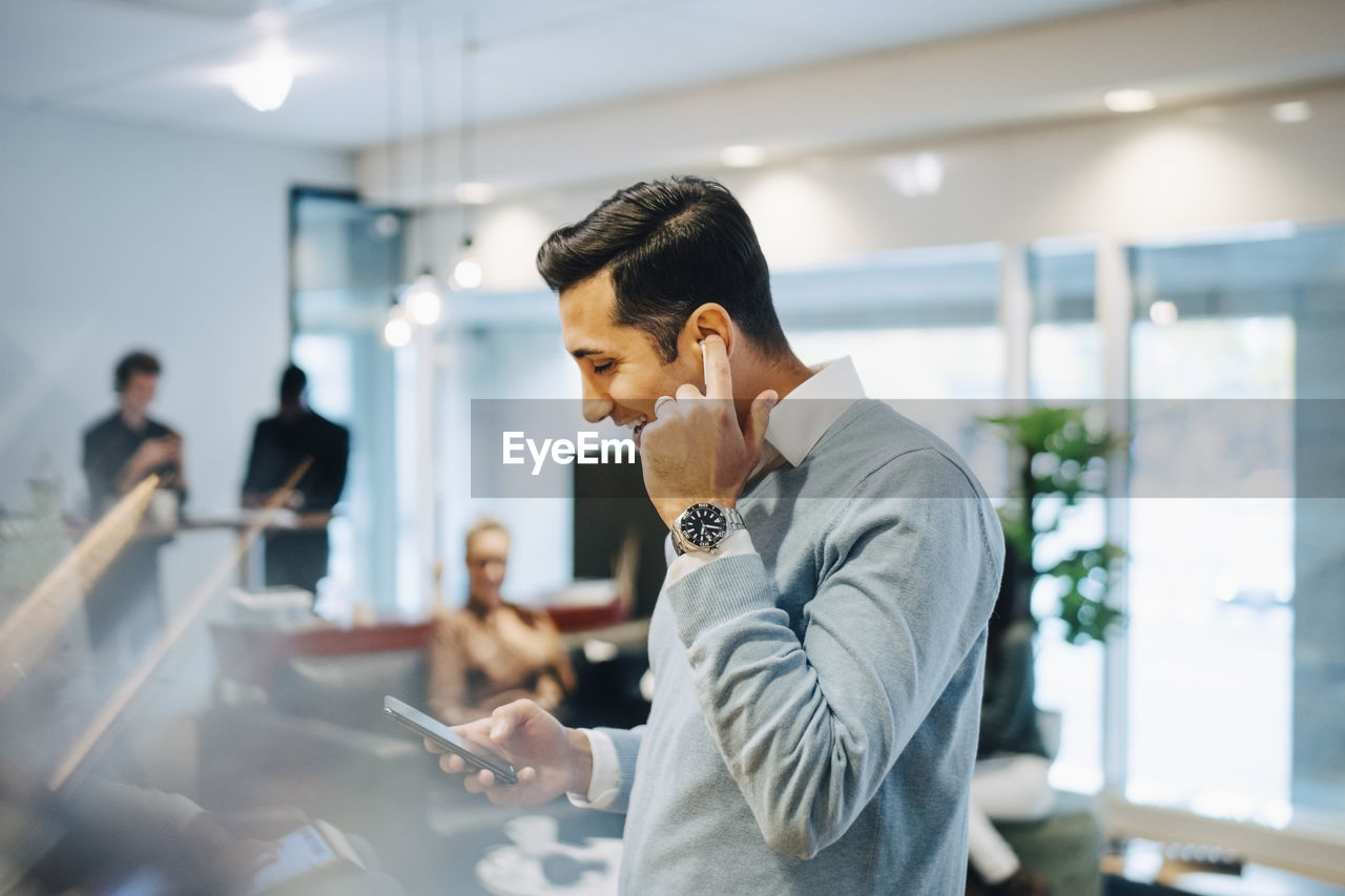 Happy businessman using smart phone while adjusting earphones in creative office