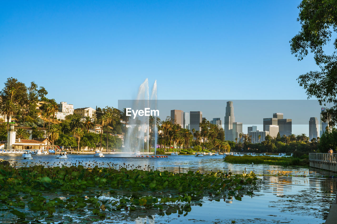 Fountain by lake and buildings against clear sky at echo park