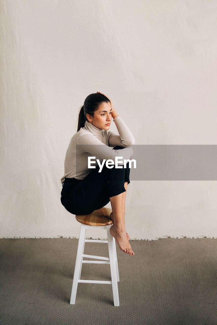 Side view of young unhappy barefoot female in casual clothes sitting on stool and looking away