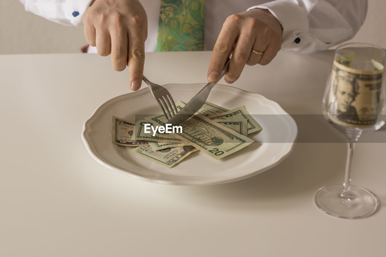 Midsection of man cutting paper currencies in plate with fork and table knife