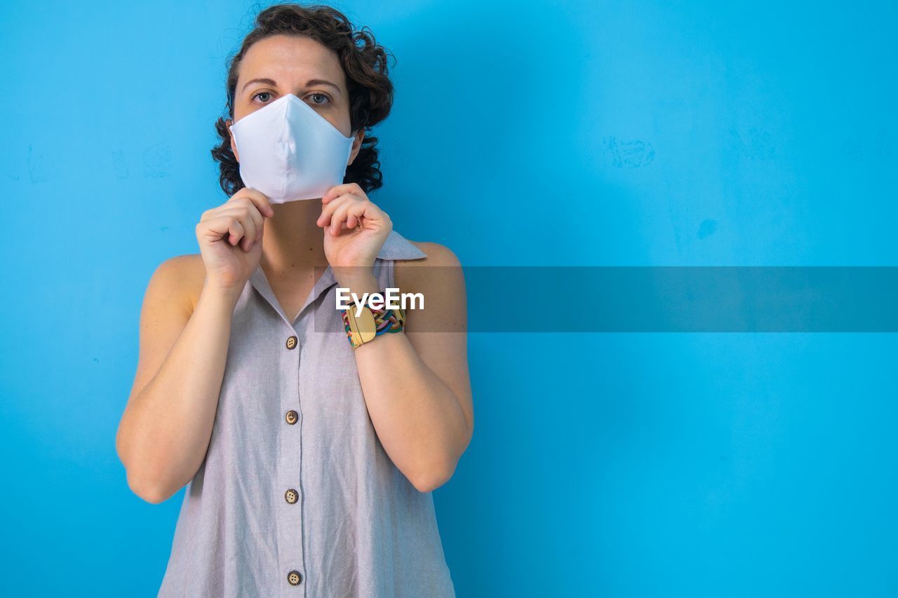 Woman on blue background preparing to take off her mask after the pandemic with copy space