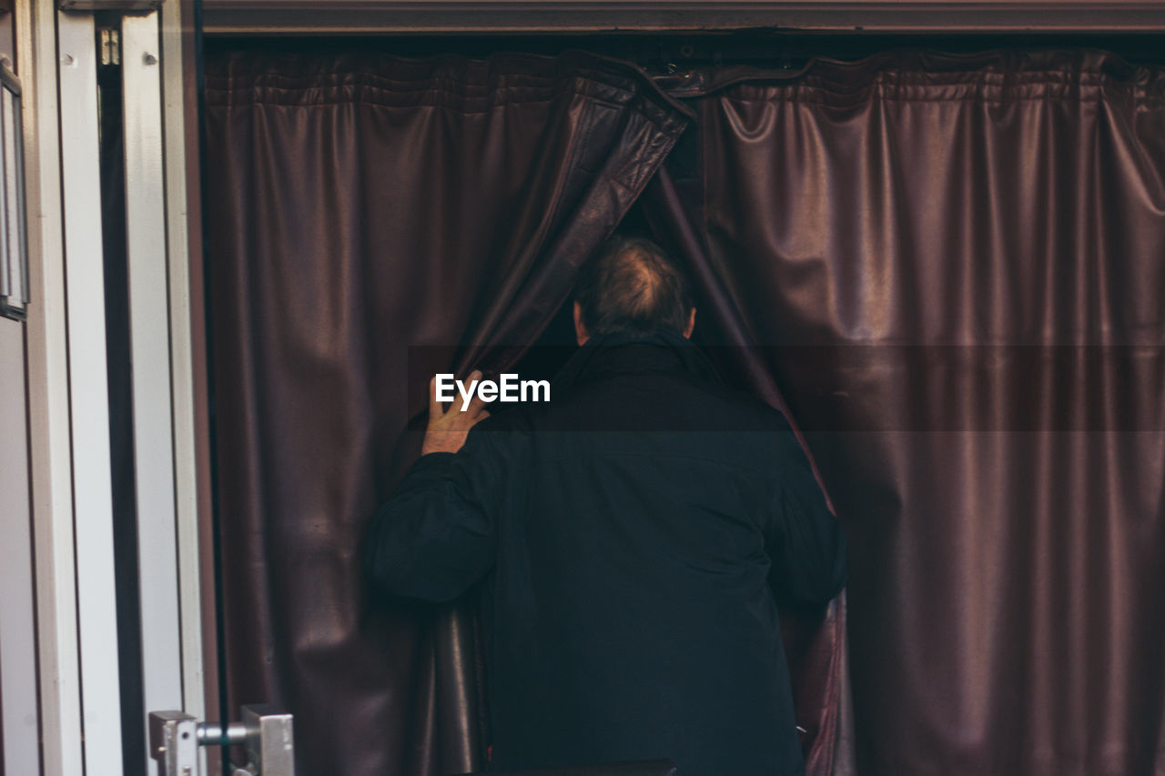 REAR VIEW OF MAN STANDING BY CURTAIN