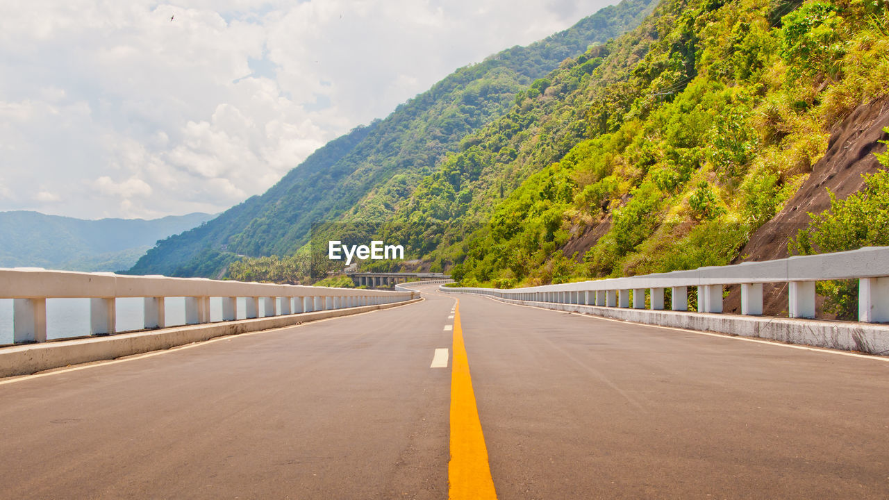 Empty road by mountains against cloudy sky