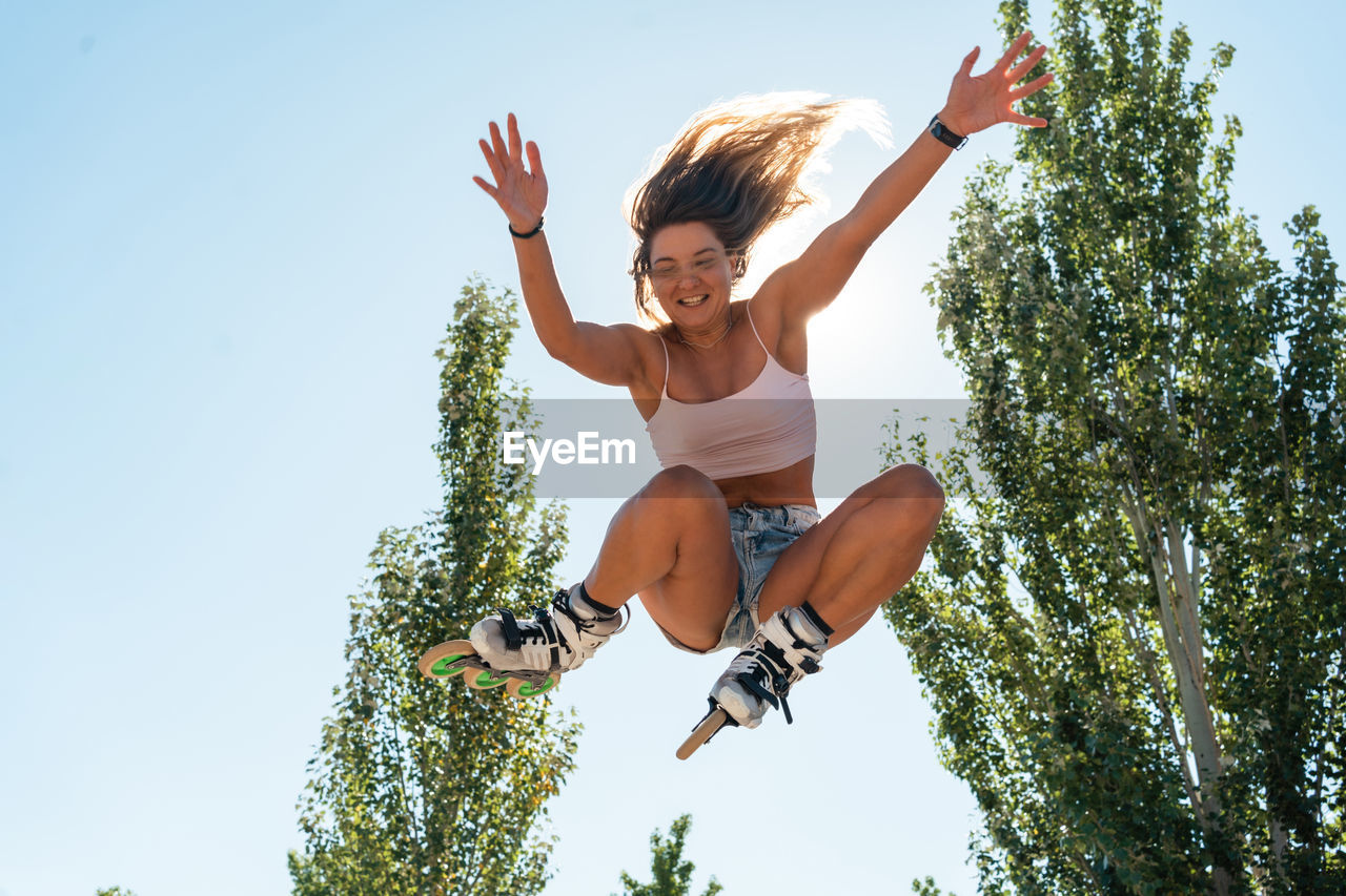 From below of active female in rollerblades jumping and performing trick in park against blue sky in summer on sunny day