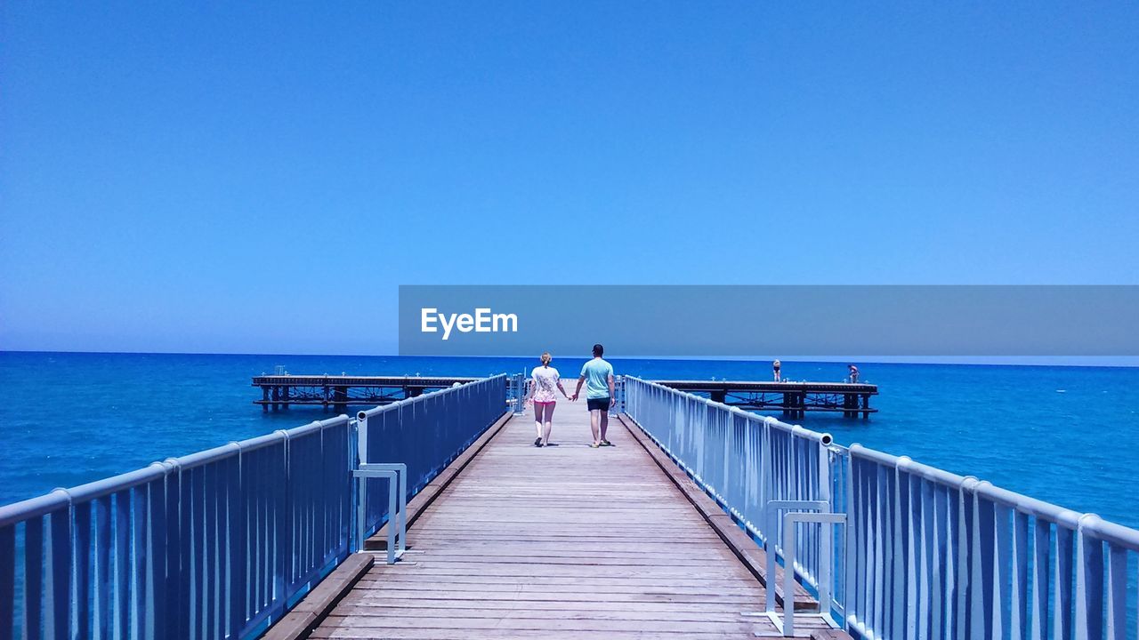 Rear view of people walking on pier at sea against clear sky