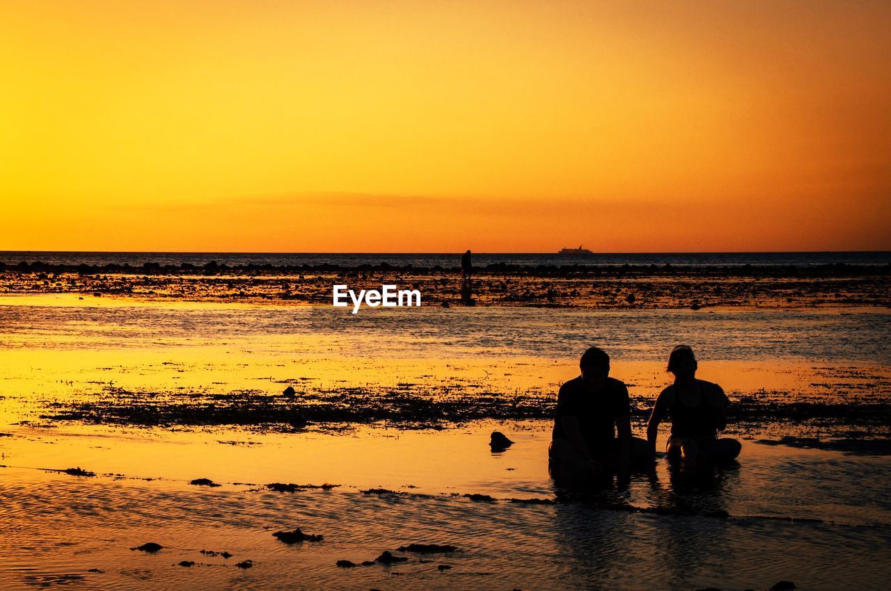 Silhouette men sitting on beach against sky during sunset