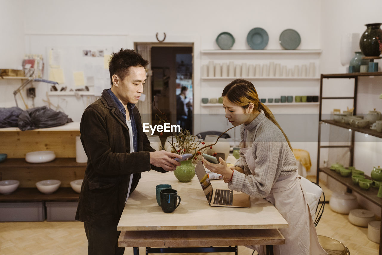 Male customer with smart phone talking to female potter while paying at workshop