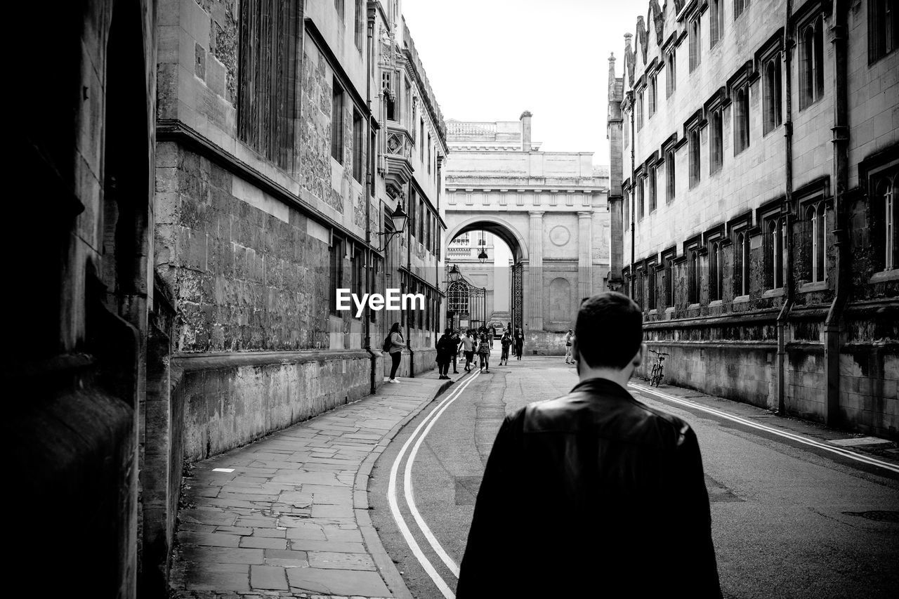 REAR VIEW OF MAN WALKING ON STREET AGAINST BUILDINGS IN CITY