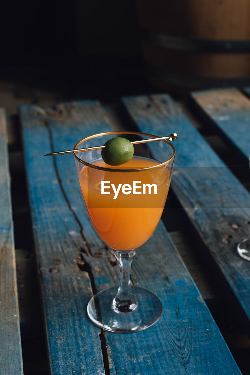 Carrot orange drink in wine glass with green olive on gold cocktail pick on blue wooden pallet 