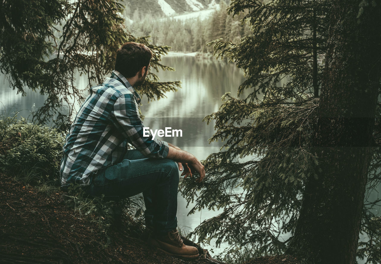 Side view of young man looking at lake while sitting in forest