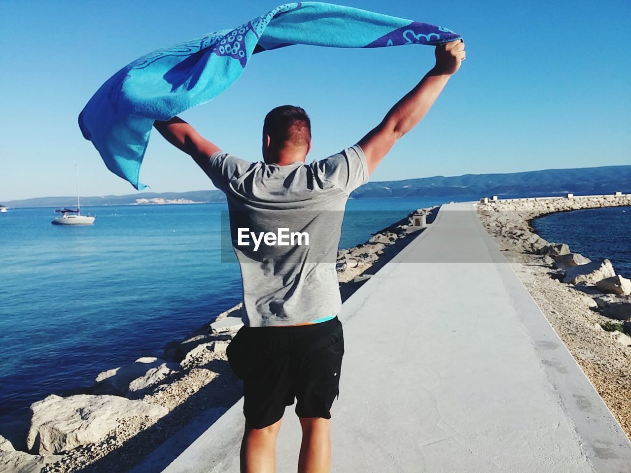 Rear view of man with towel standing on pier at beach against clear blue sky