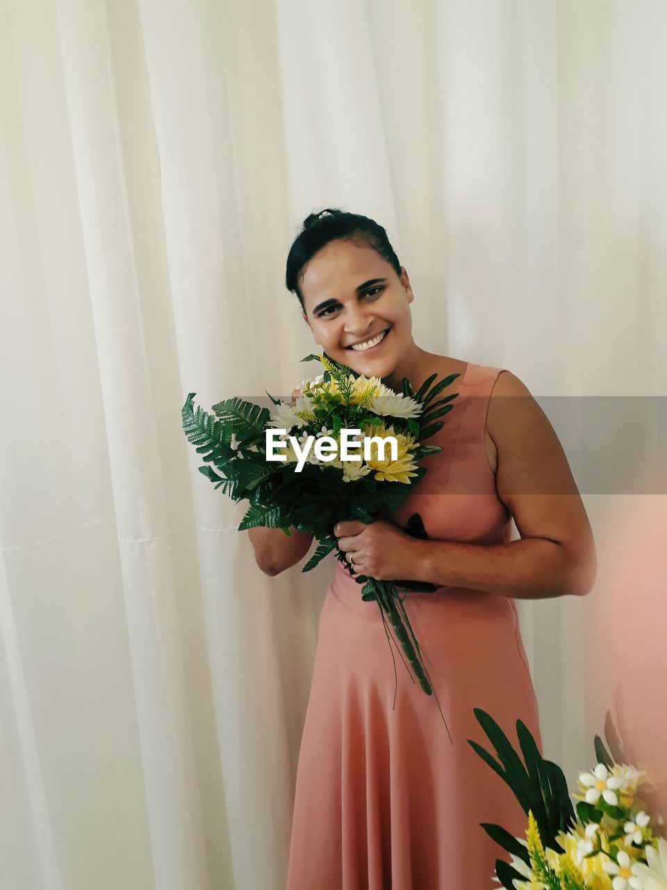 Portrait of smiling woman holding bouquet while standing against curtain