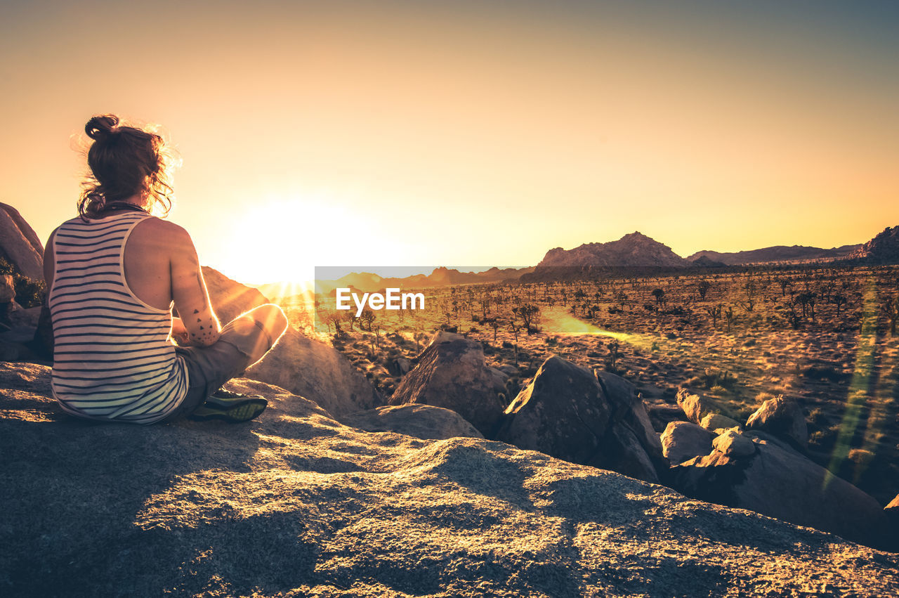 Rear view of man sitting on rock formation while looking at field against sky during sunset