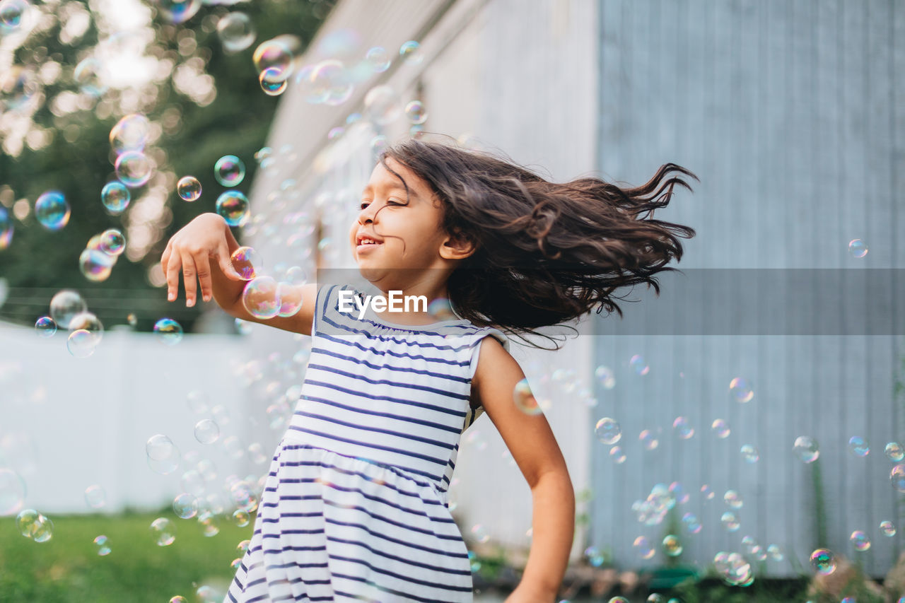 Diverse mixed race pre school girl outdoors during summer having fun in backyard with bubbles 