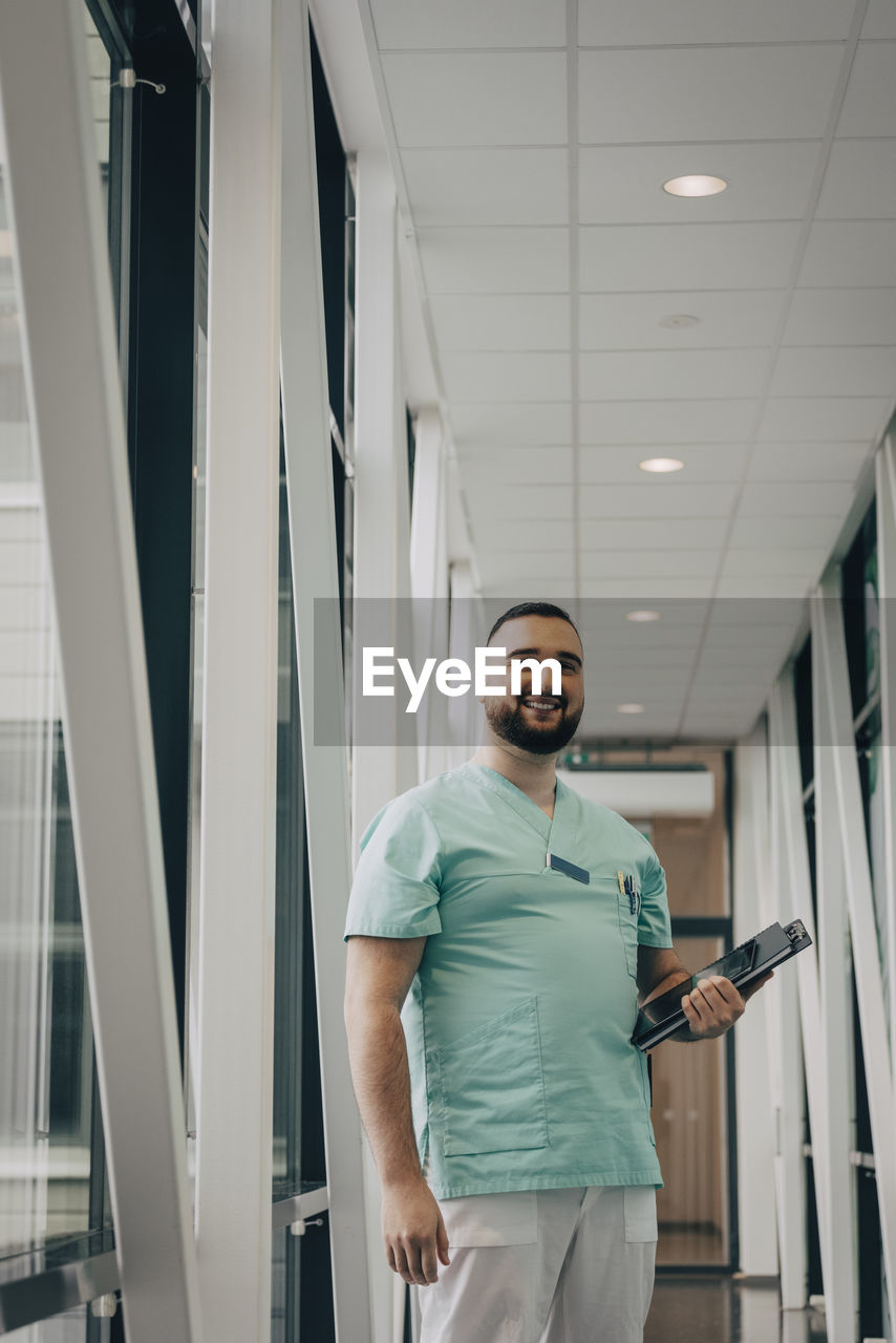 Smiling male healthcare worker holding file while standing in corridor of hospital