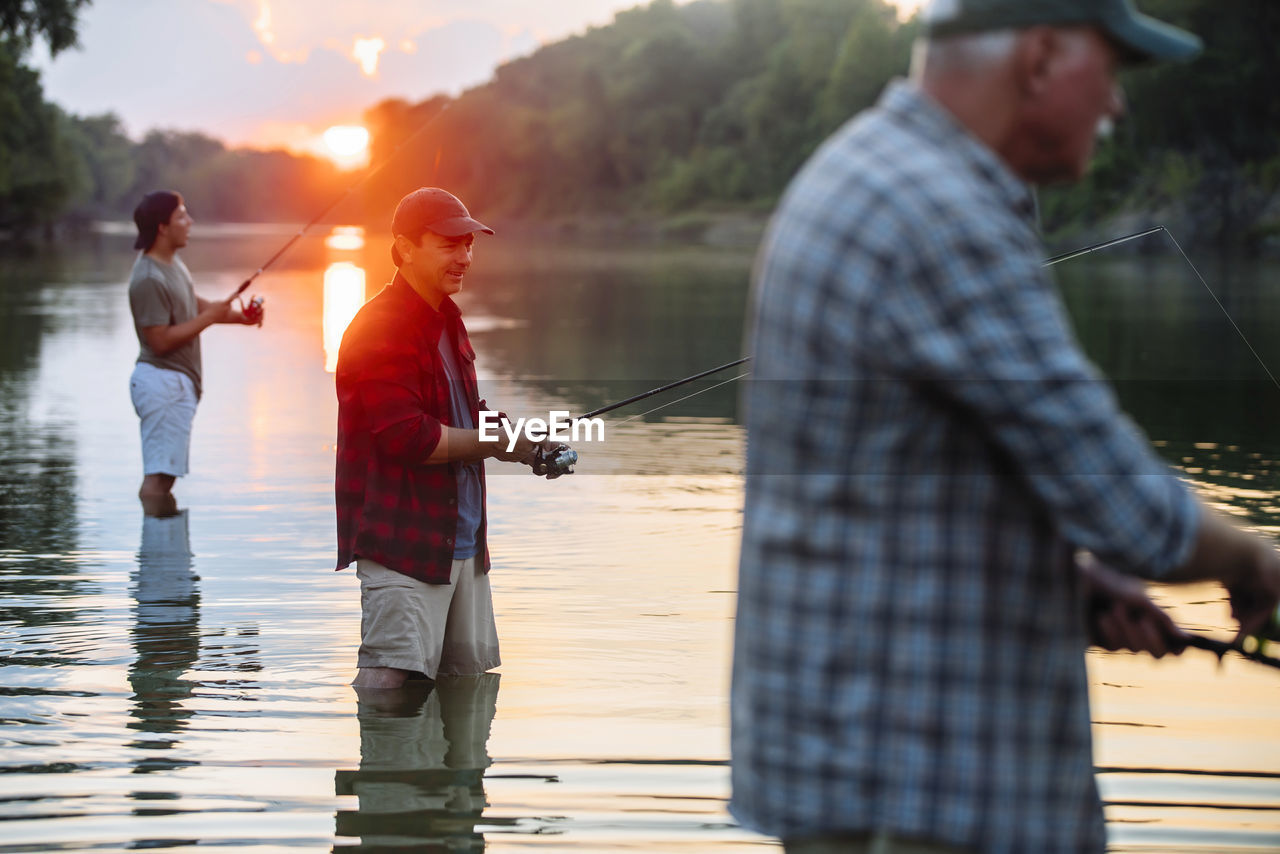 Side view of male friends fishing while standing in lake during sunset