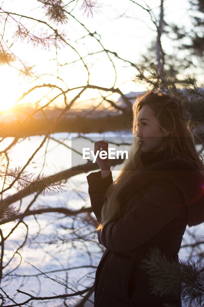 Side view of young woman in warm clothing standing by trees during sunset