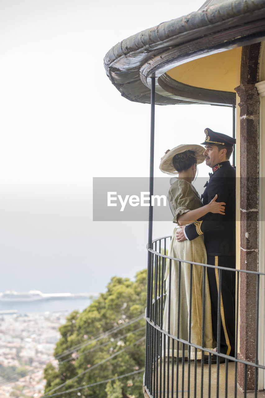 MAN AND WOMAN STANDING BY RAILING AGAINST SKY