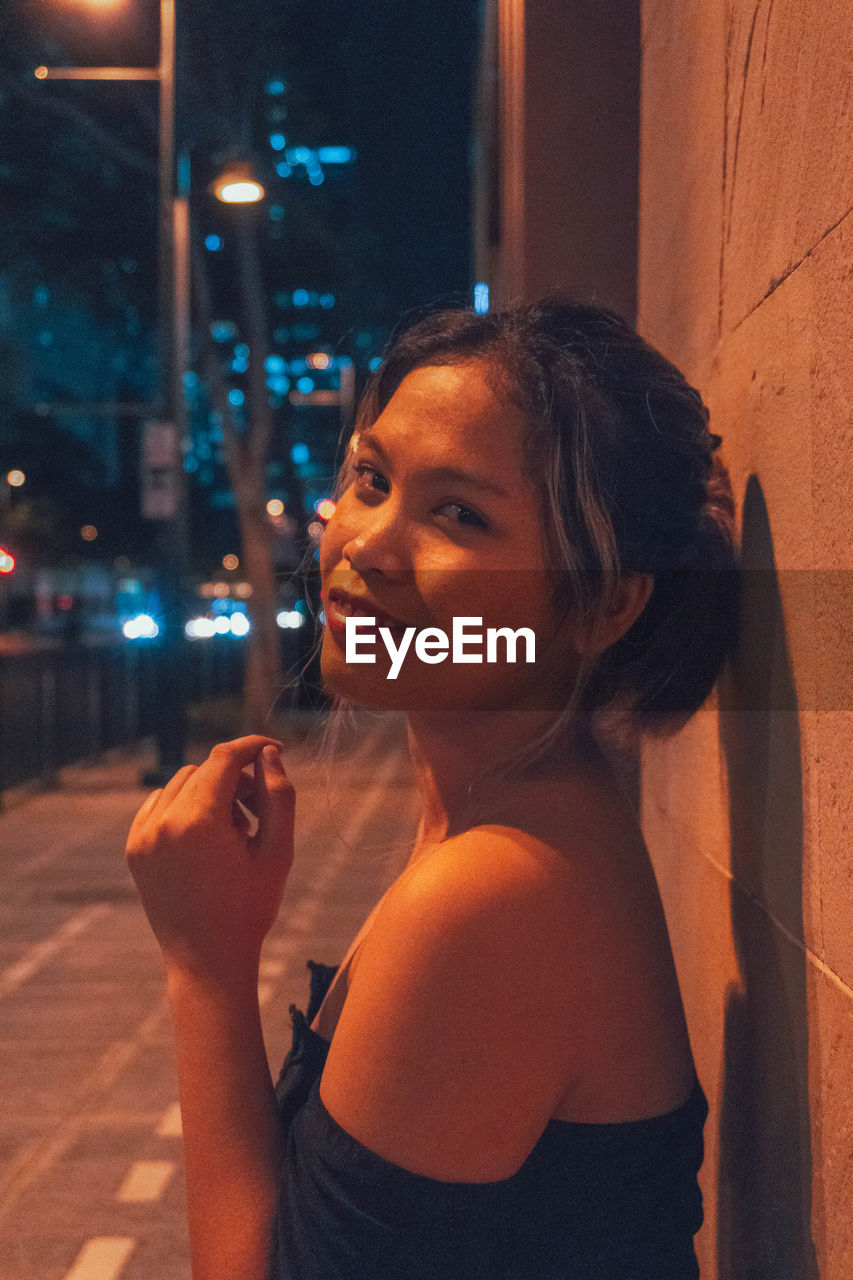 PORTRAIT OF YOUNG WOMAN STANDING BY ILLUMINATED CITY AT NIGHT