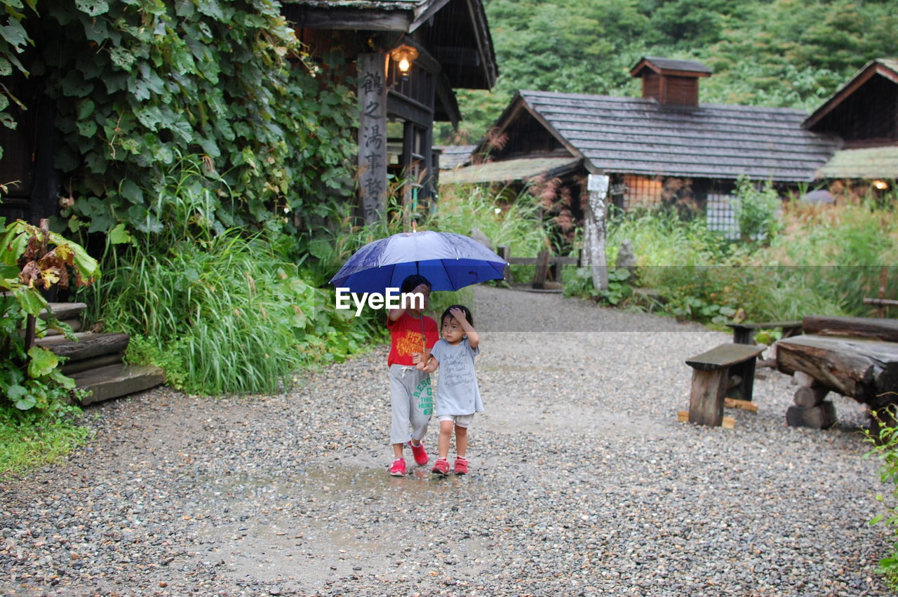 Siblings with umbrella walking on dirt road amidst houses