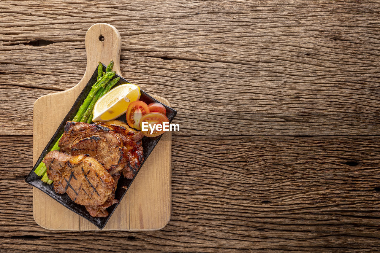 Grilled pork chop with asparagus, lemon and tomato in black ceramic plate on cutting board