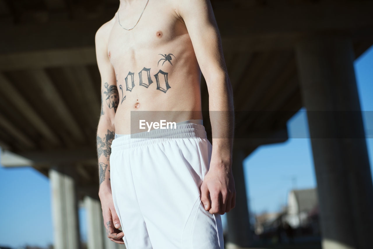 Midsection of shirtless man with tattoo standing outdoors