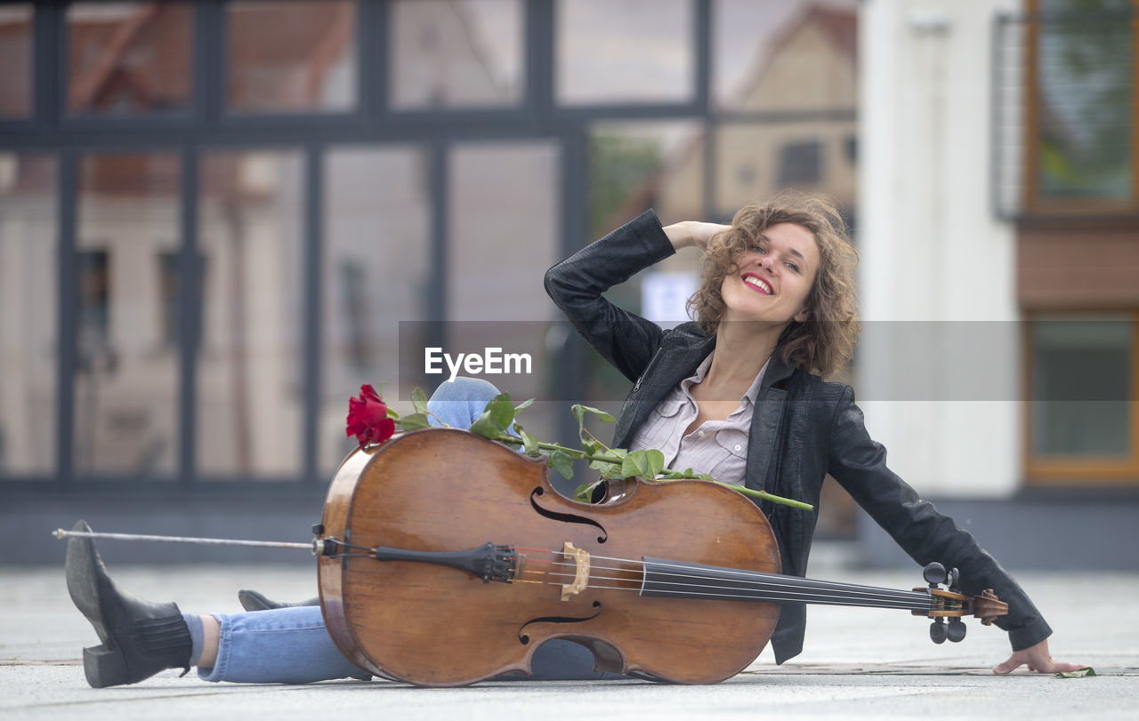 one person, adult, women, music, young adult, arts culture and entertainment, musical instrument, string instrument, city, musician, sitting, architecture, smiling, happiness, long hair, full length, hairstyle, emotion, lifestyles, cello, blond hair, day, clothing, enjoyment, city life, cheerful, casual clothing, leisure activity, portrait, person, nature, musical equipment, outdoors, individuality, female, performance, holding, front view, street