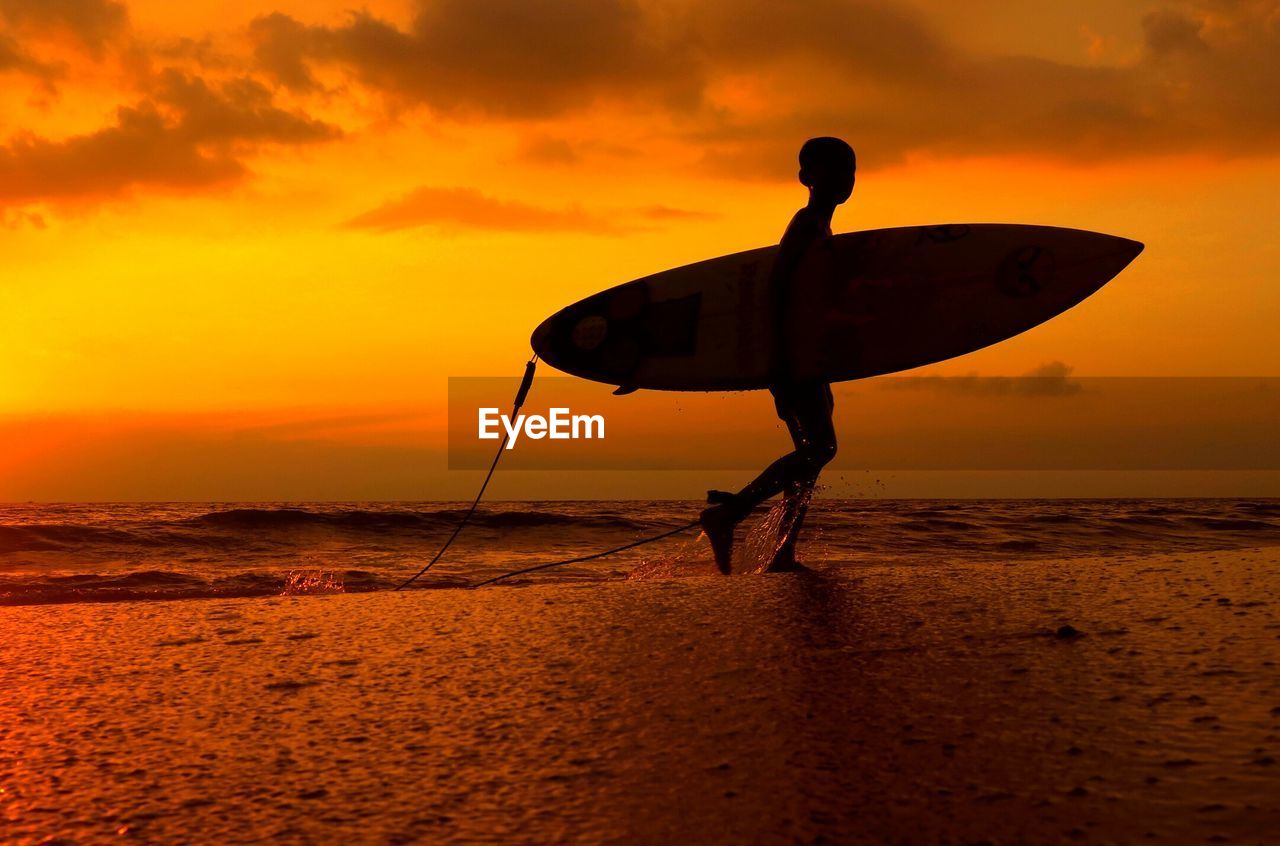 Silhouette boy carrying surfboard on shore at beach against orange sky during sunset