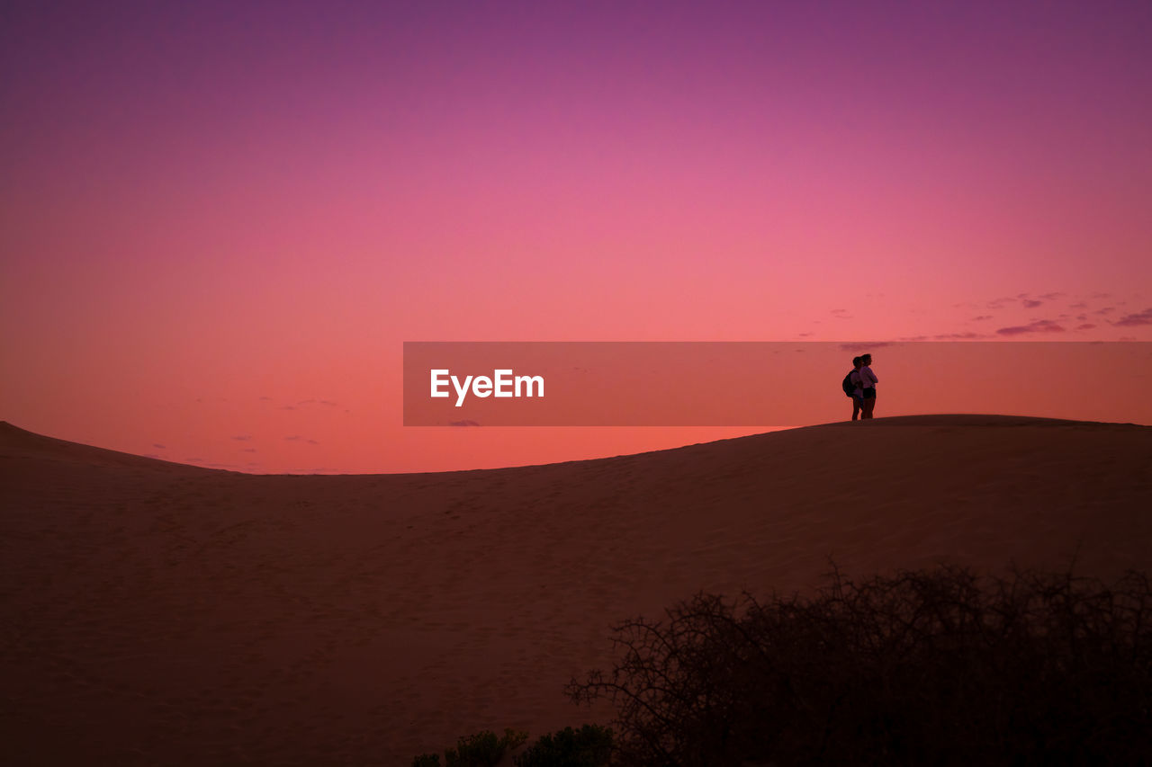 Silhouette friends standing on desert against sky during sunset