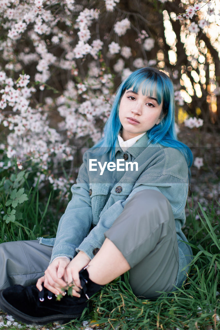 Sad millennial female model with blue hair in stylish outfit looking at camera thoughtfully while sitting on green grass near blooming tree in spring garden