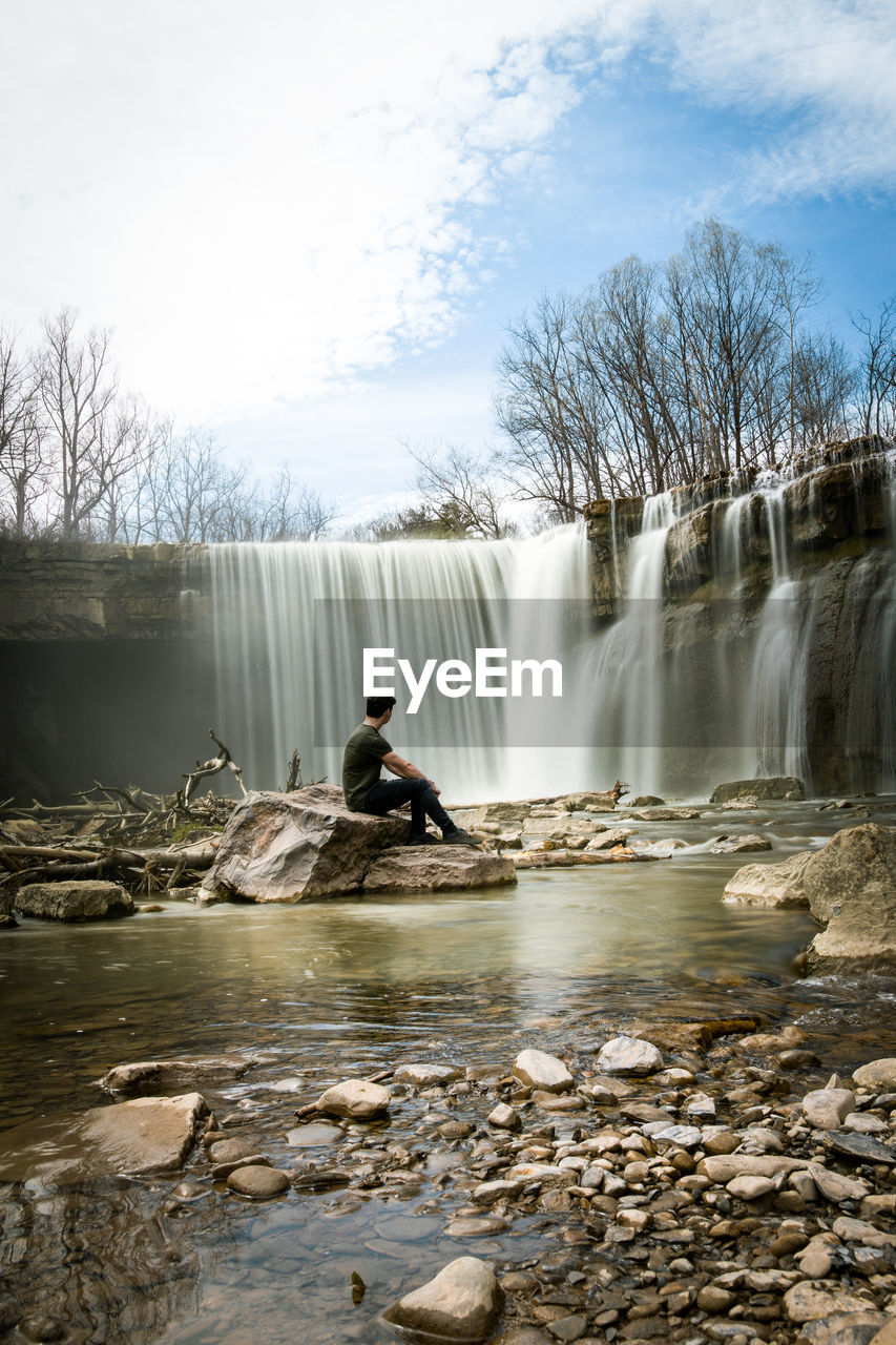 Man sitting by waterfall against sky