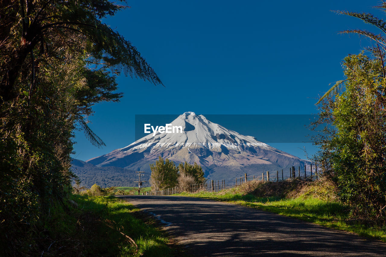 Empty road leading towards snowcapped mountain against clear blue sky