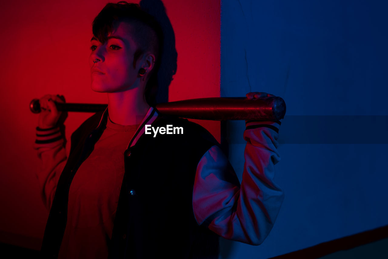 Unconventional woman with modern hairstyle standing against wall with baseball bat behind head and looking away illuminated by red and blue at city street