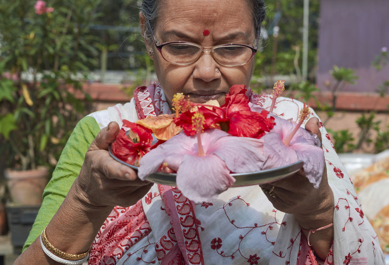 An aged bengali woman offering prayer to sun god, holding bouquet of hibiscus flowers in tray.
