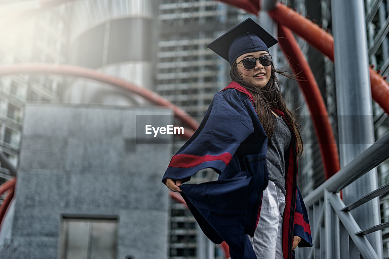 Low angle view portrait of young woman wearing graduation gown while standing against building