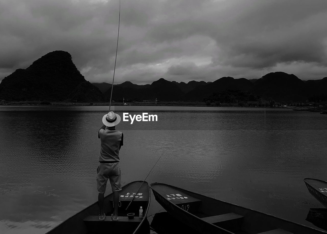 Rear view of man standing on boat while fishing in lake during sunset