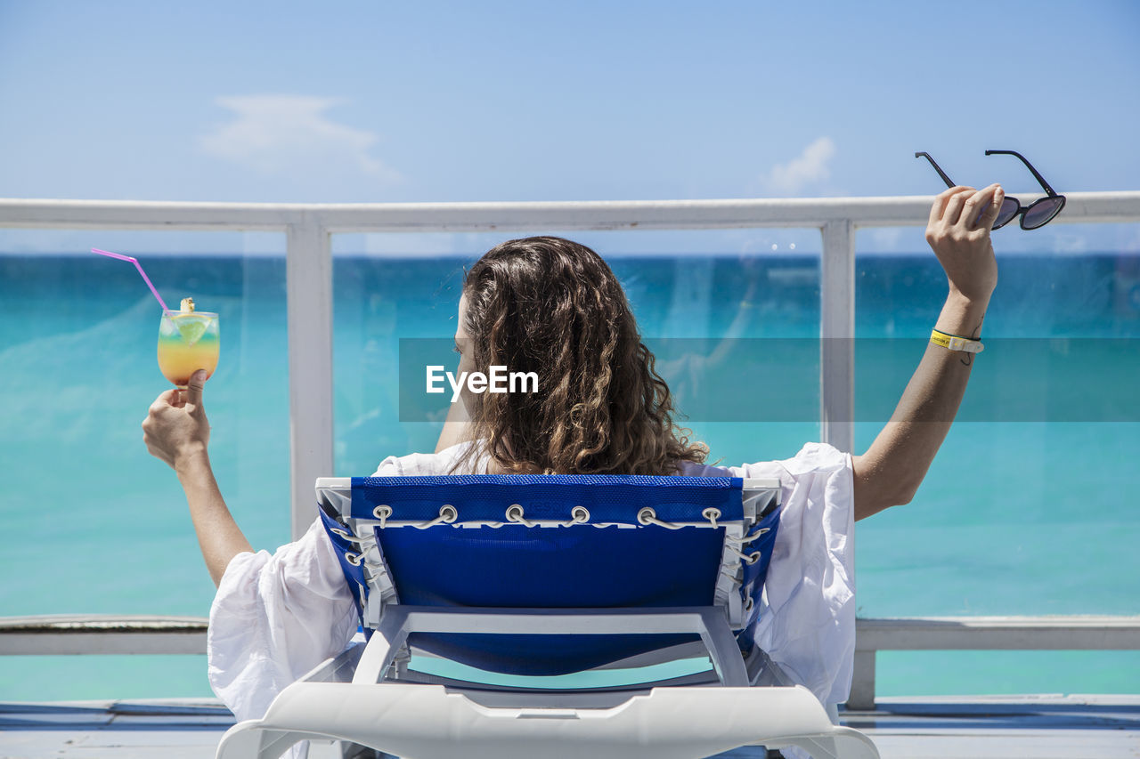 Rear view of woman having drink while relaxing on lounge chair against sea