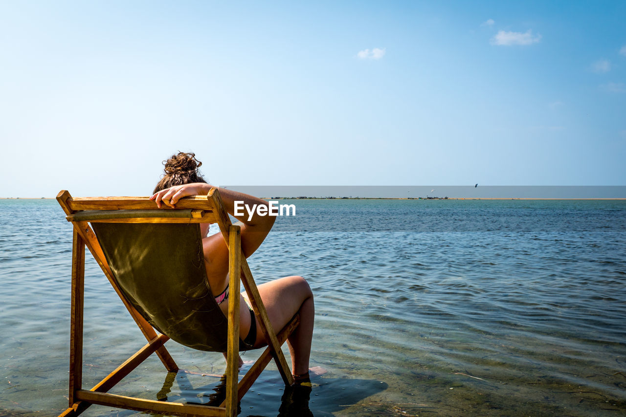 Rear view of young woman sitting on deck chair by sea against sky