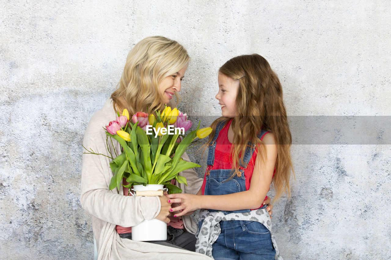 Smiling mother and daughter with bouquet standing against wall
