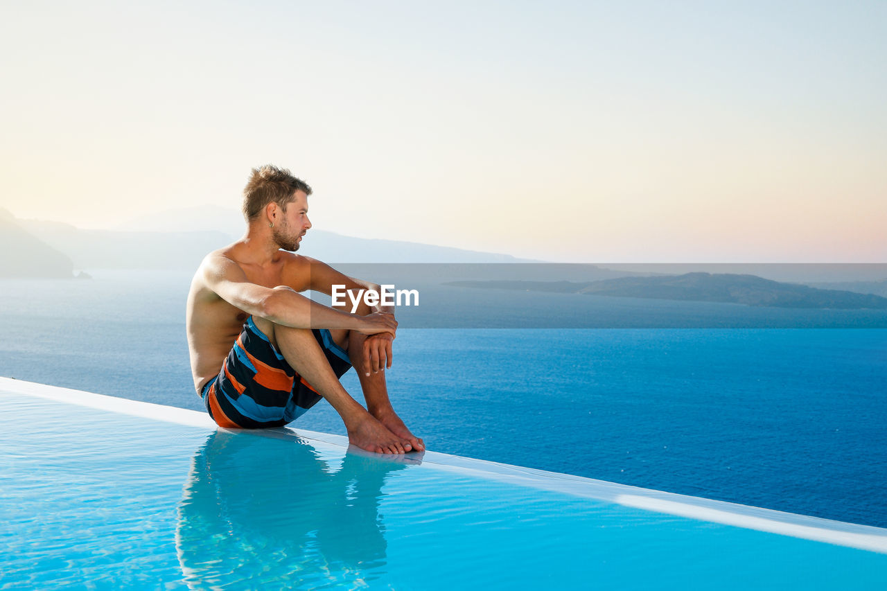 Young man sitting at poolside against sea