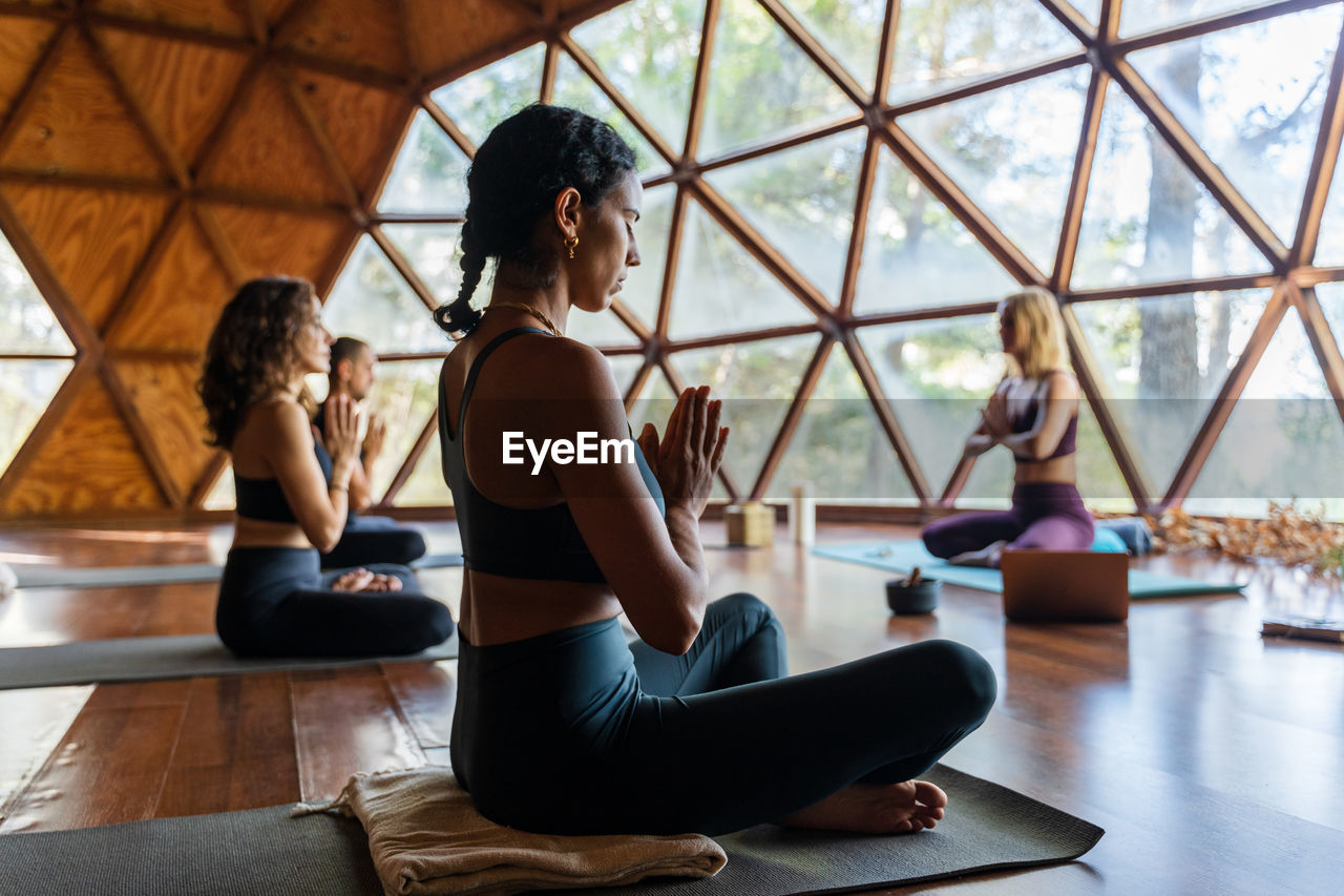 People in front of her teacher in sportswear while are seating in lotus pose with namaste hands while practicing breathing exercise during yoga session in studio