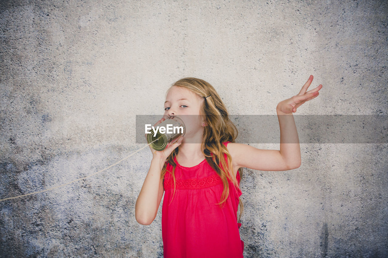 Cute girl playing with tin can phone while standing against wall